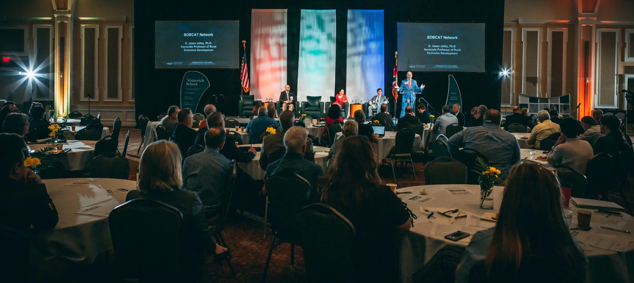 A wide-angle view of a large conference hall during a BOBCAT Network presentation. Dr. G. Jason Jolley is speaking on stage in front of a large audience. The stage features banners for the Voinovich School of Leadership and Public Service.