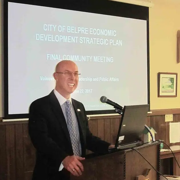 A presenter, a man in a black suit, standing at a wooden podium with a microphone and laptop, smiles while gesturing toward a large presentation screen behind him. The screen reads, "CITY OF BELPRE ECONOMIC DEVELOPMENT STRATEGIC PLAN FINAL COMMUNITY MEETING," with smaller text below, "Voinovich School of Leadership and Public Affairs" and the date "June 22, 2017."