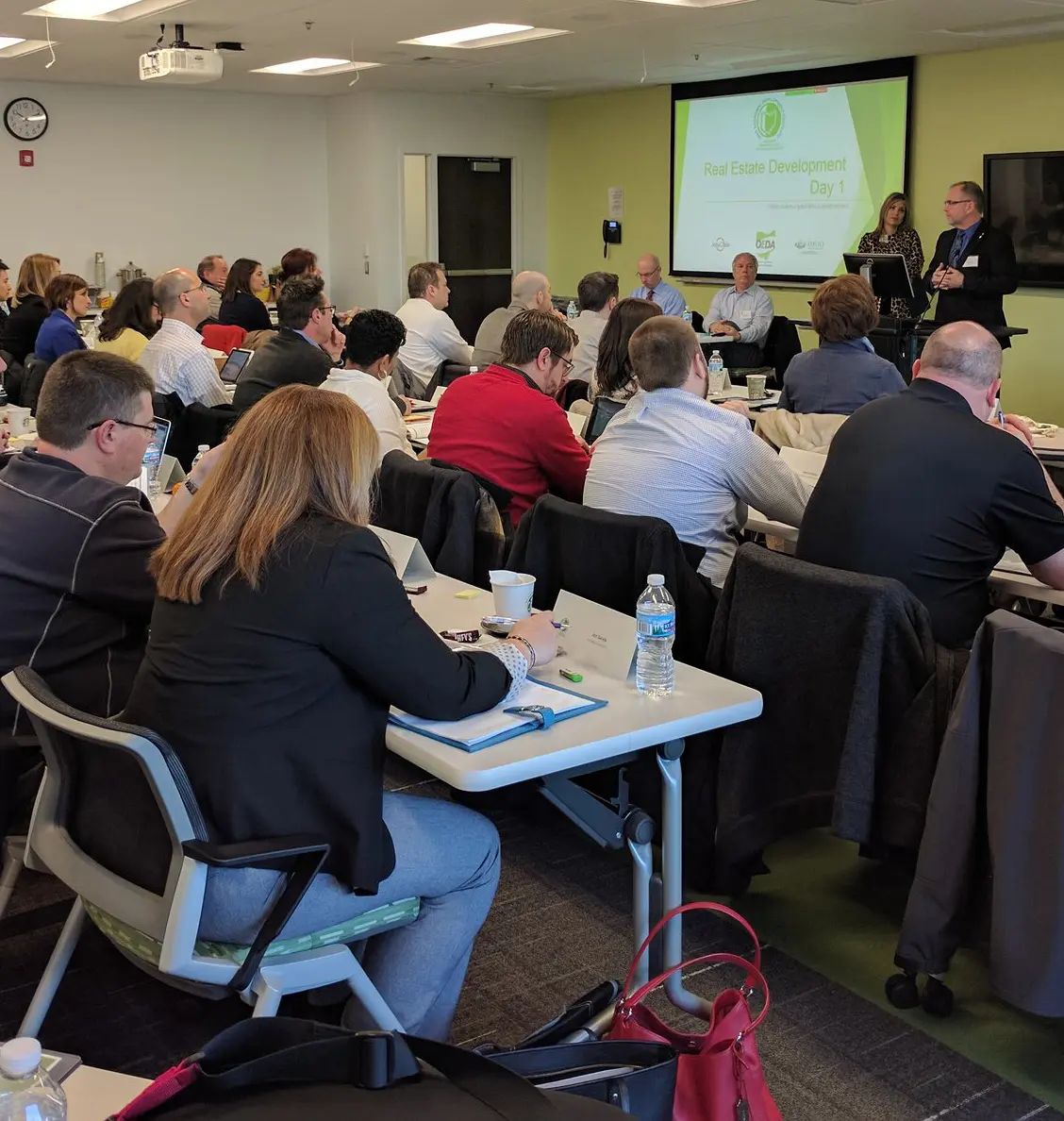 A classroom setting at Ohio University where a group of professionals is attending a 'Real Estate Development Day 1' workshop. Two presenters are speaking at the front near a projection screen showing the course title and partner logos.